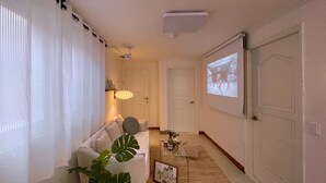 A living room with a sofa, a coffee table, a potted plant, and a wall-mounted projector displaying a scene. - House Lotus (Seoul)