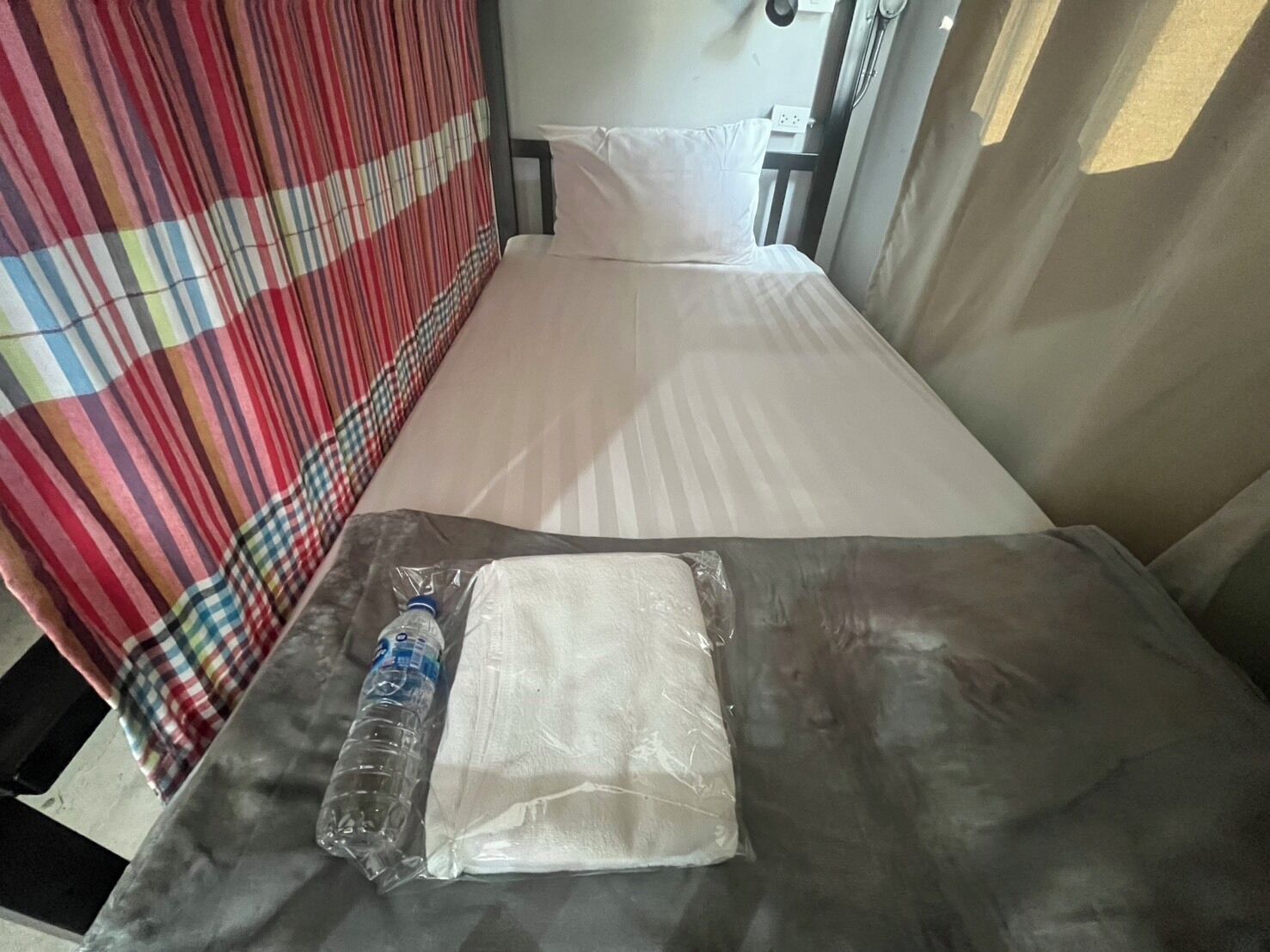 Une petite chambre avec un lit simple, un rideau à rayures, une bouteille d’eau et un vêtement blanc plié sur le lit.