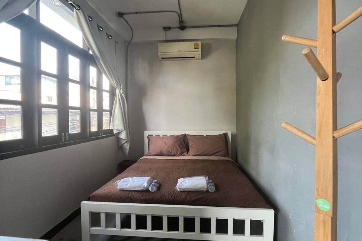 A compact room with a single bed, a small table, a window with curtains, an air conditioning unit, and a wooden rack.