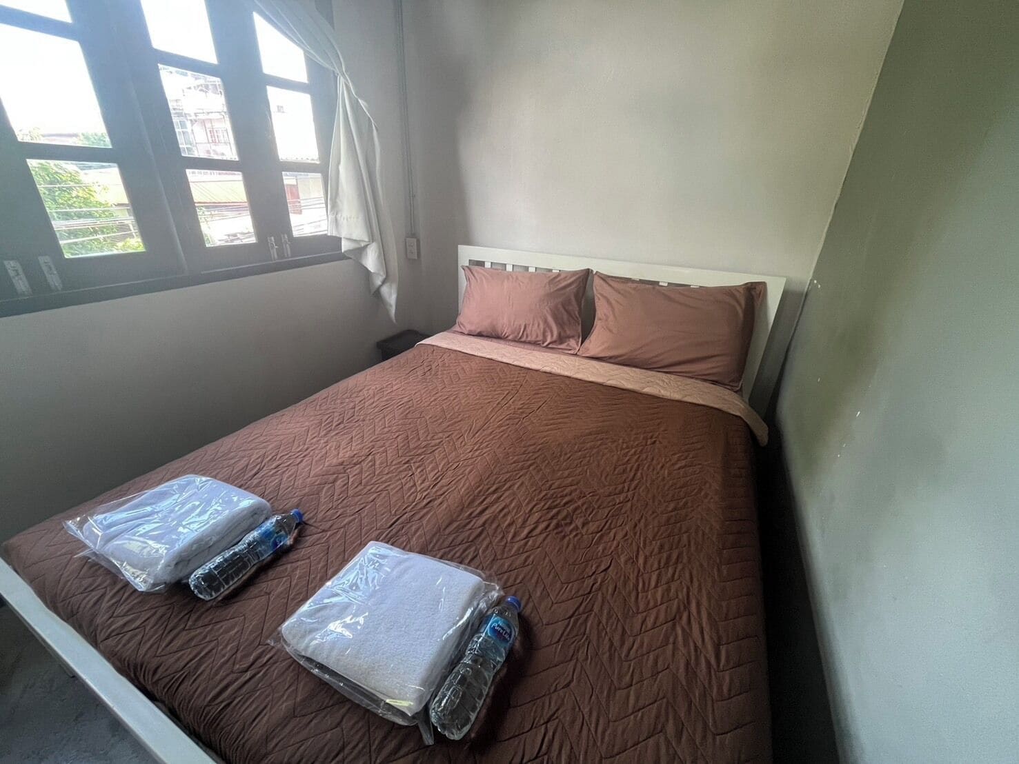 A single bed with a brown quilt, two pillows, and two plastic-wrapped items on the bed.
