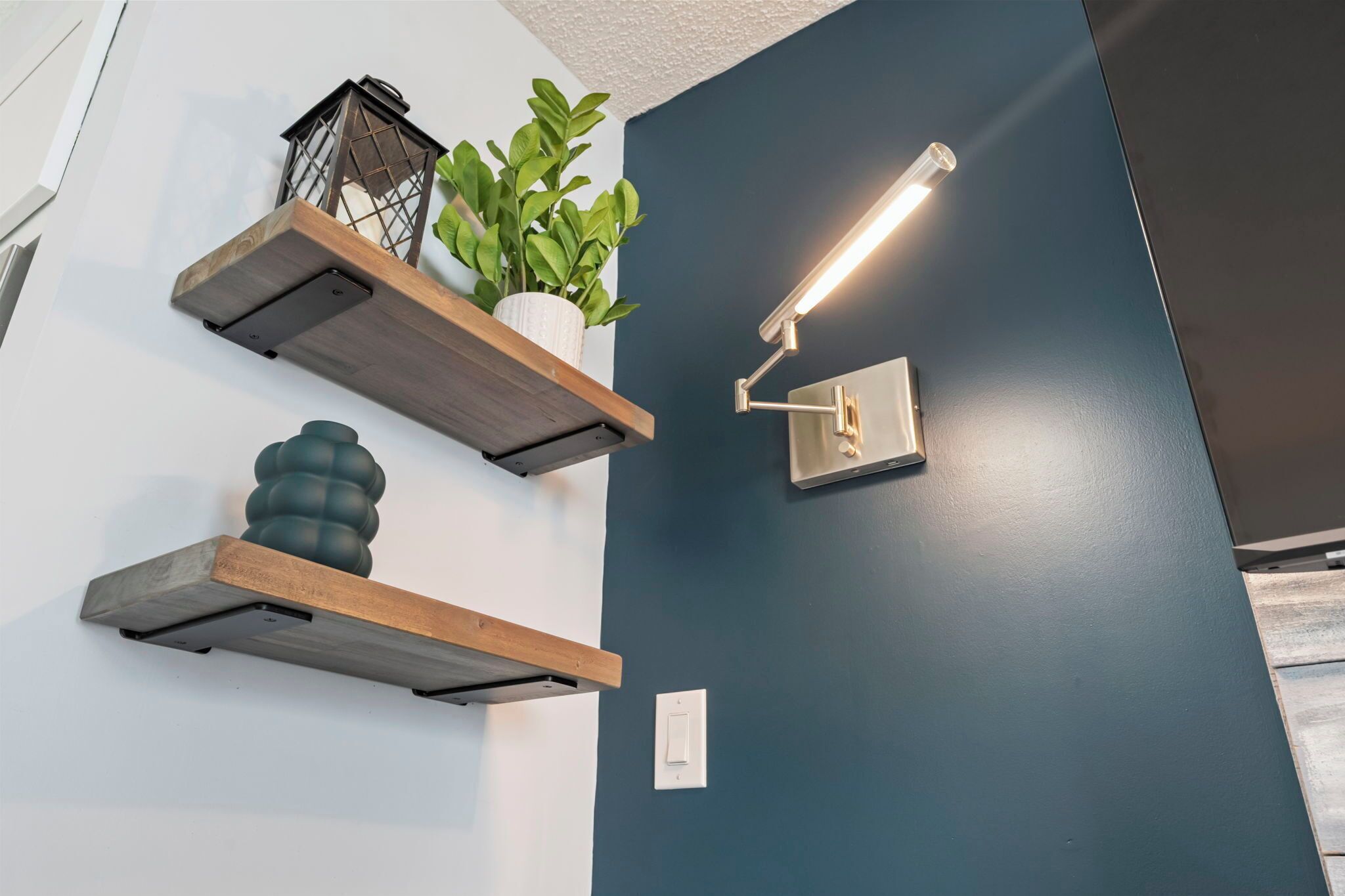 A room with a dark blue wall, wooden floating shelves, a potted plant, a wall-mounted light fixture, and a switch.