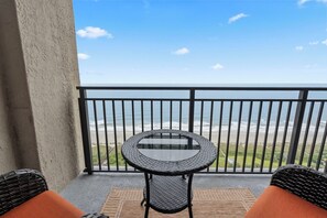 Een balkon met een ronde tafel en twee rieten stoelen, met uitzicht op een strand.