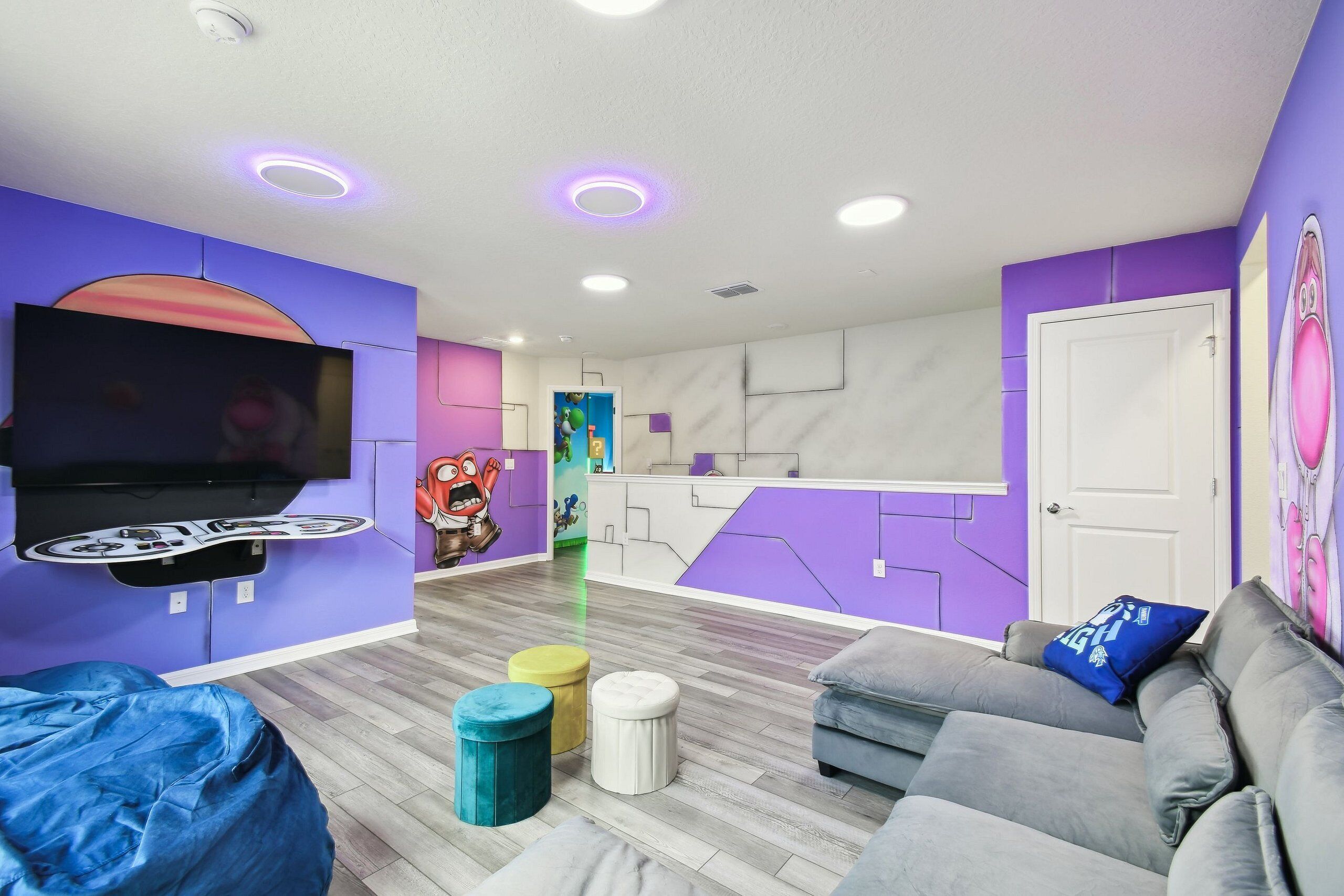 Un moderno salón con televisión de pantalla plana montada en la pared, un sofá, otomanas y un mural colorido.