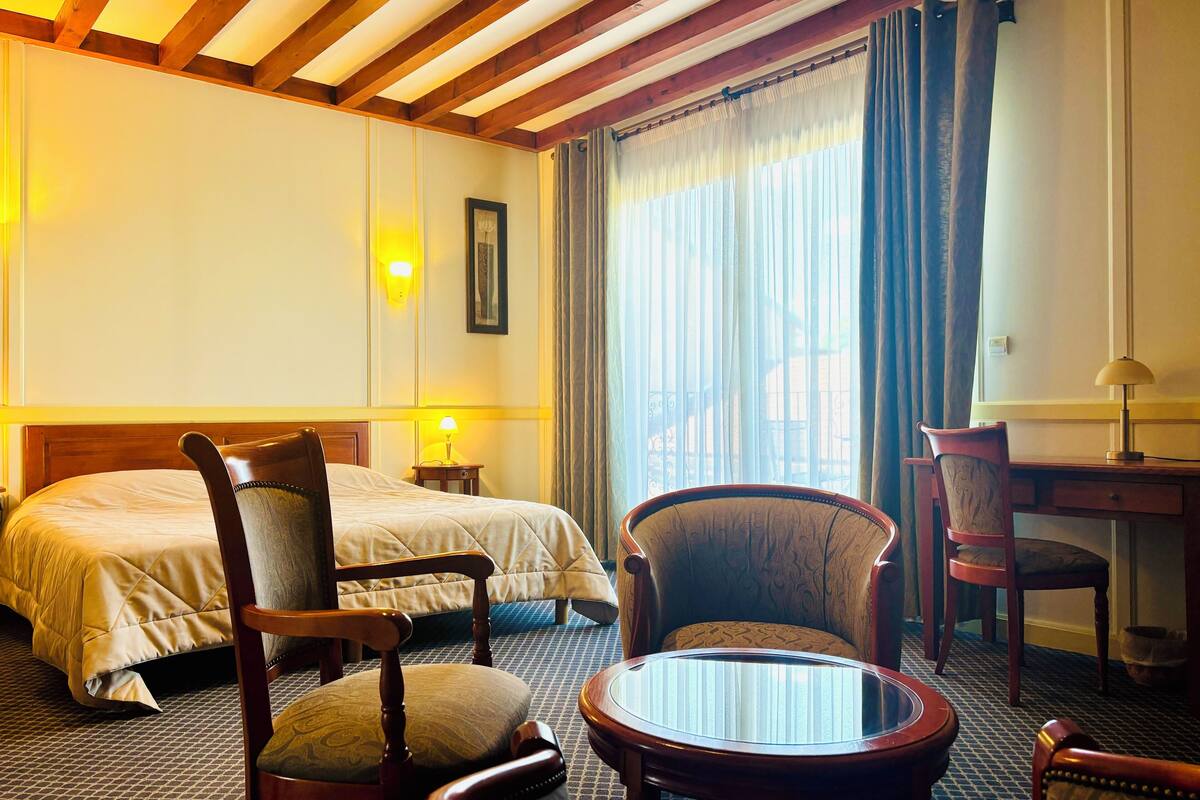 A hotel room with a bed, two armchairs, a small round table, a desk, and a window with curtains.