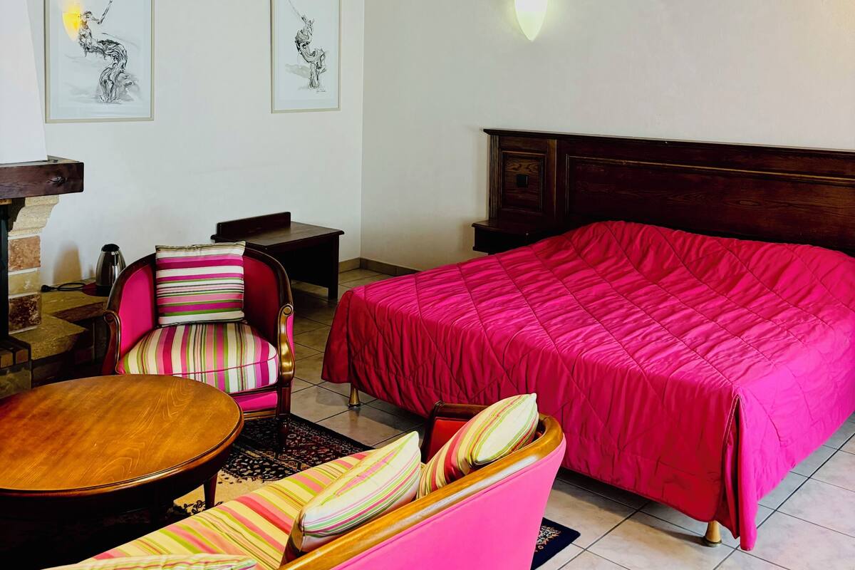 A room with a wooden ceiling, a bed with a red bedspread, a pink armchair, a wooden coffee table, and two framed artworks on the wall.