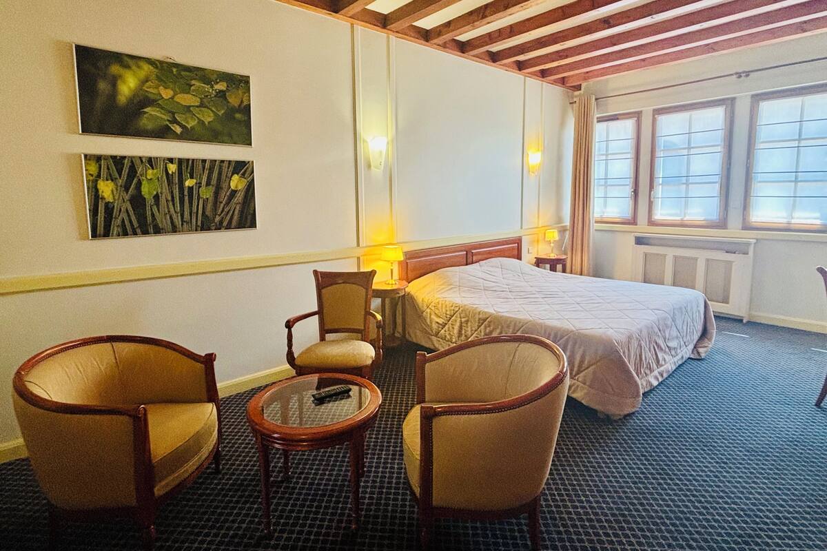 A hotel room with a bed, two armchairs, a small table, a lamp, and artwork on the wall.