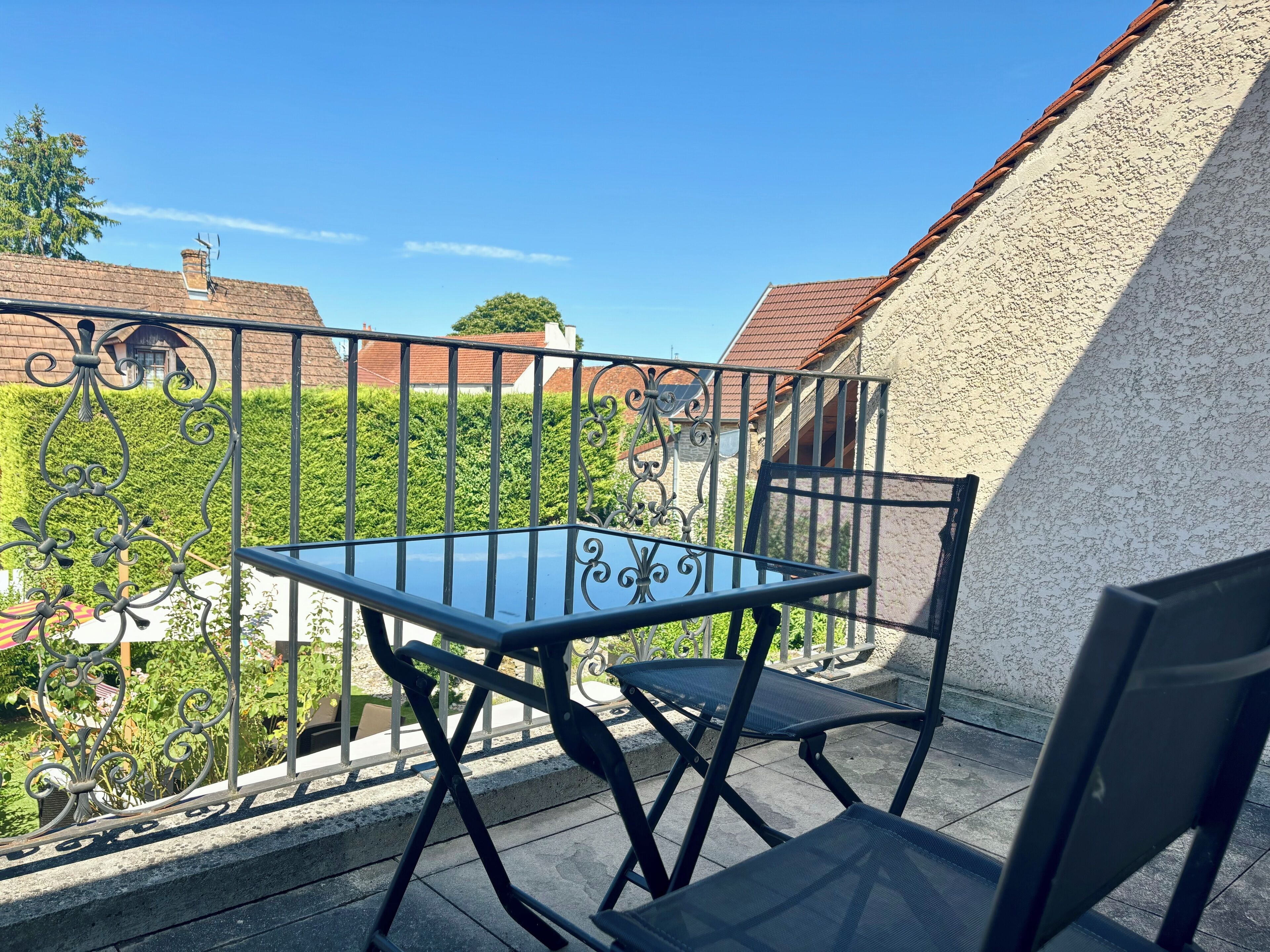 A balcony with a glass table and two chairs, overlooking a garden and houses.