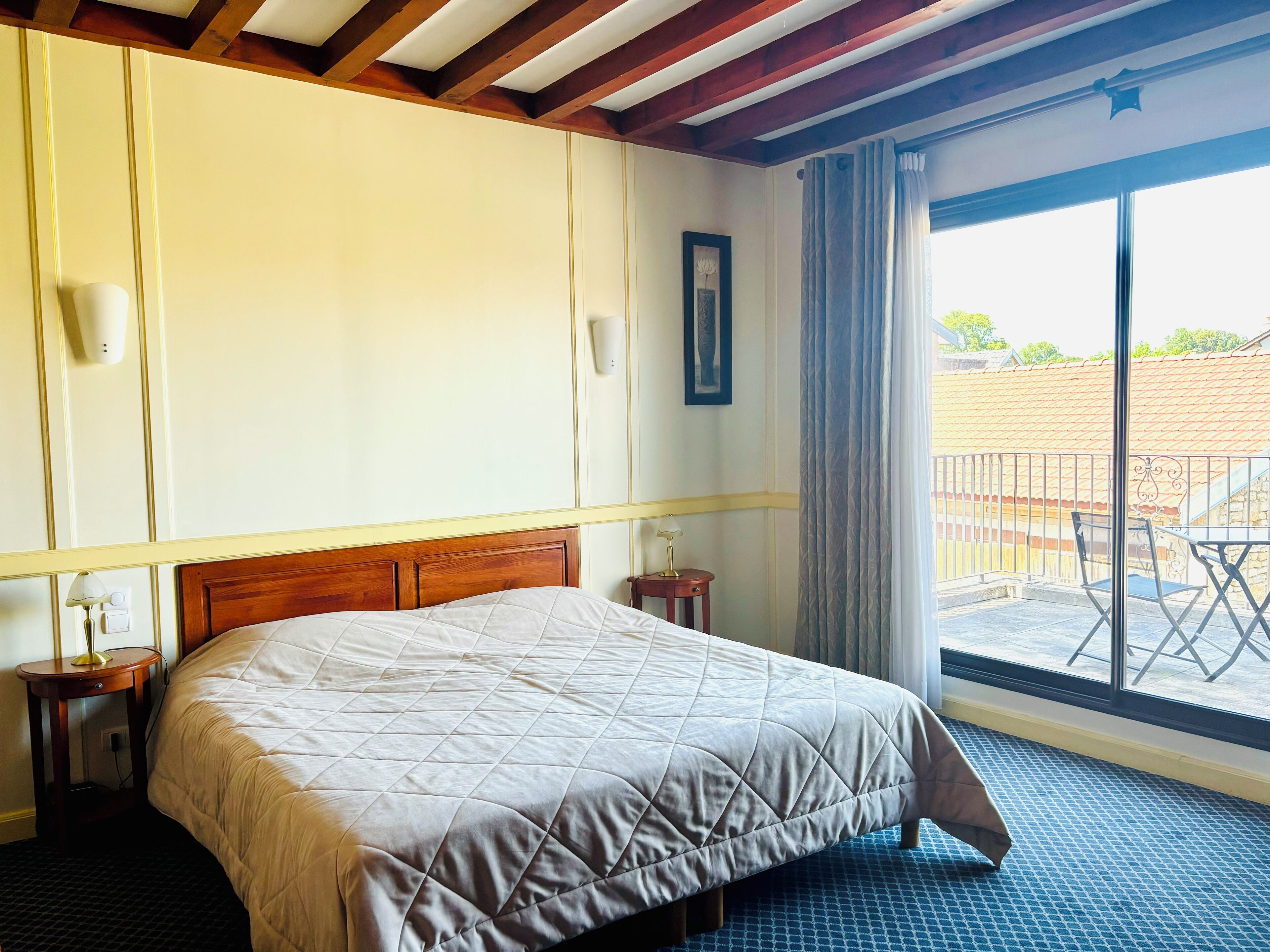 Habitación de hotel con una cama grande, un cabecero de madera, una mesita de noche pequeña con una lámpara, una luz de pared y vistas al balcón exterior.
