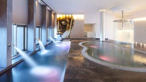 Ett modernt spaområde med en stor, rund bubbelpool, en massagestol som är monterad på väggen och en rad fönster längs den vänstra sidan.
