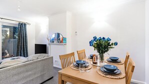 Un comedor con una mesa de madera dispuesta para cuatro, rodeada de sillas. Un jarrón con flores azules está sobre la mesa, y hay un televisor sobre un mueble contra la pared.