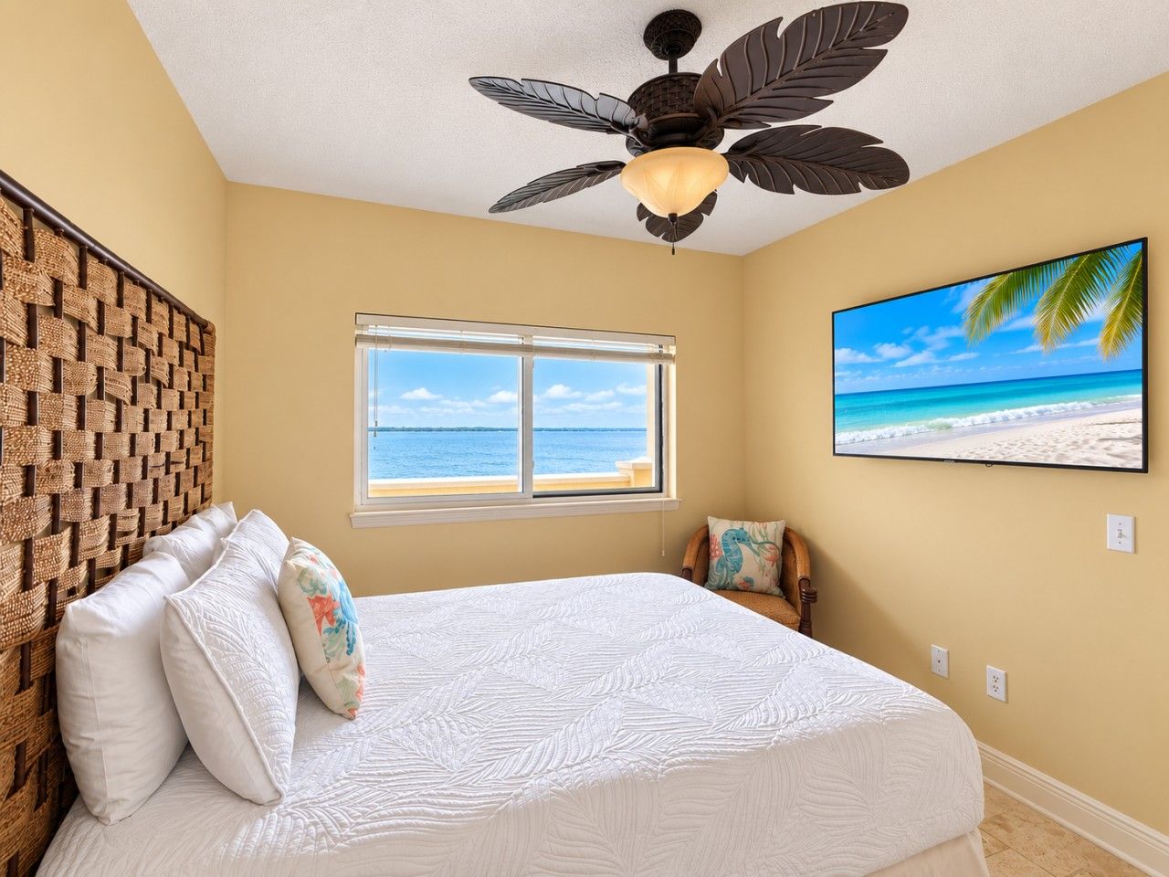 A bedroom with a bed, a ceiling fan, a window with a view of the beach, and a wall-mounted TV.