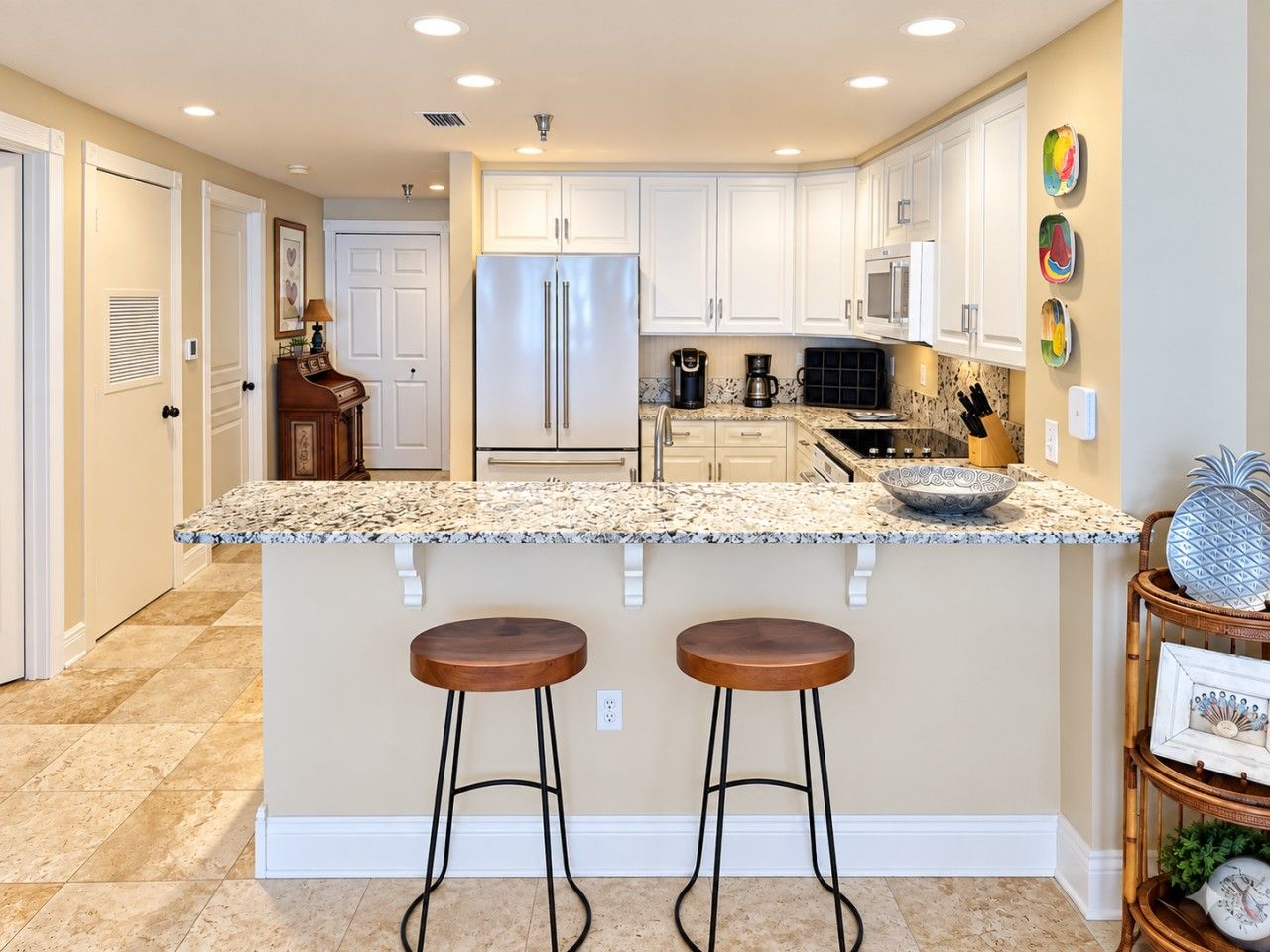 A kitchen with white cabinets, a granite countertop, two bar stools, and a refrigerator.