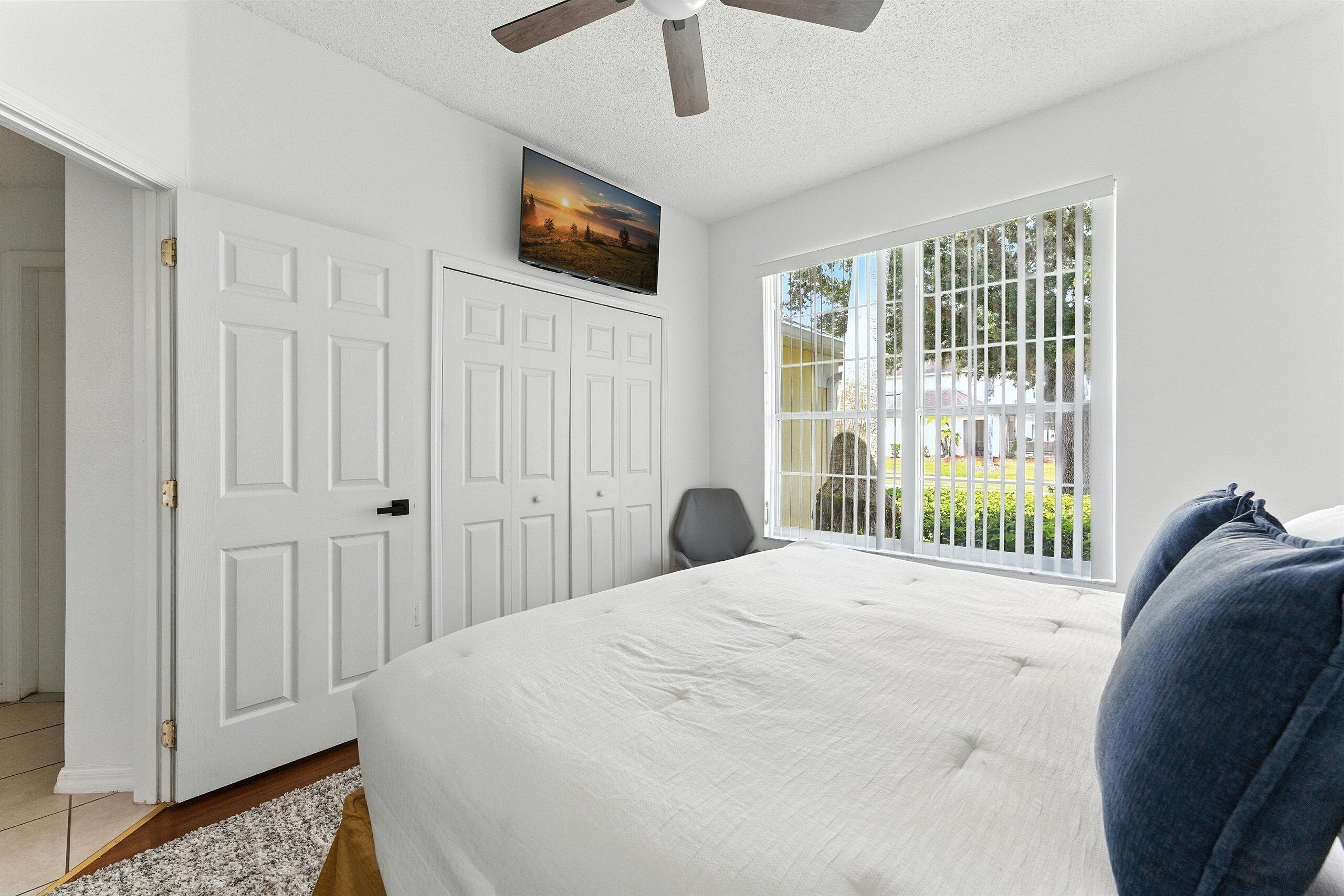 A bedroom with a large bed, a ceiling fan, a TV on the wall, and a window with a view of greenery.