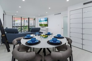 Uma zona de refeições moderna com uma mesa redonda posta para seis pessoas, rodeada por cadeiras estofadas em cinzento. Uma janela ampla oferece vista para um edifício com varandas.