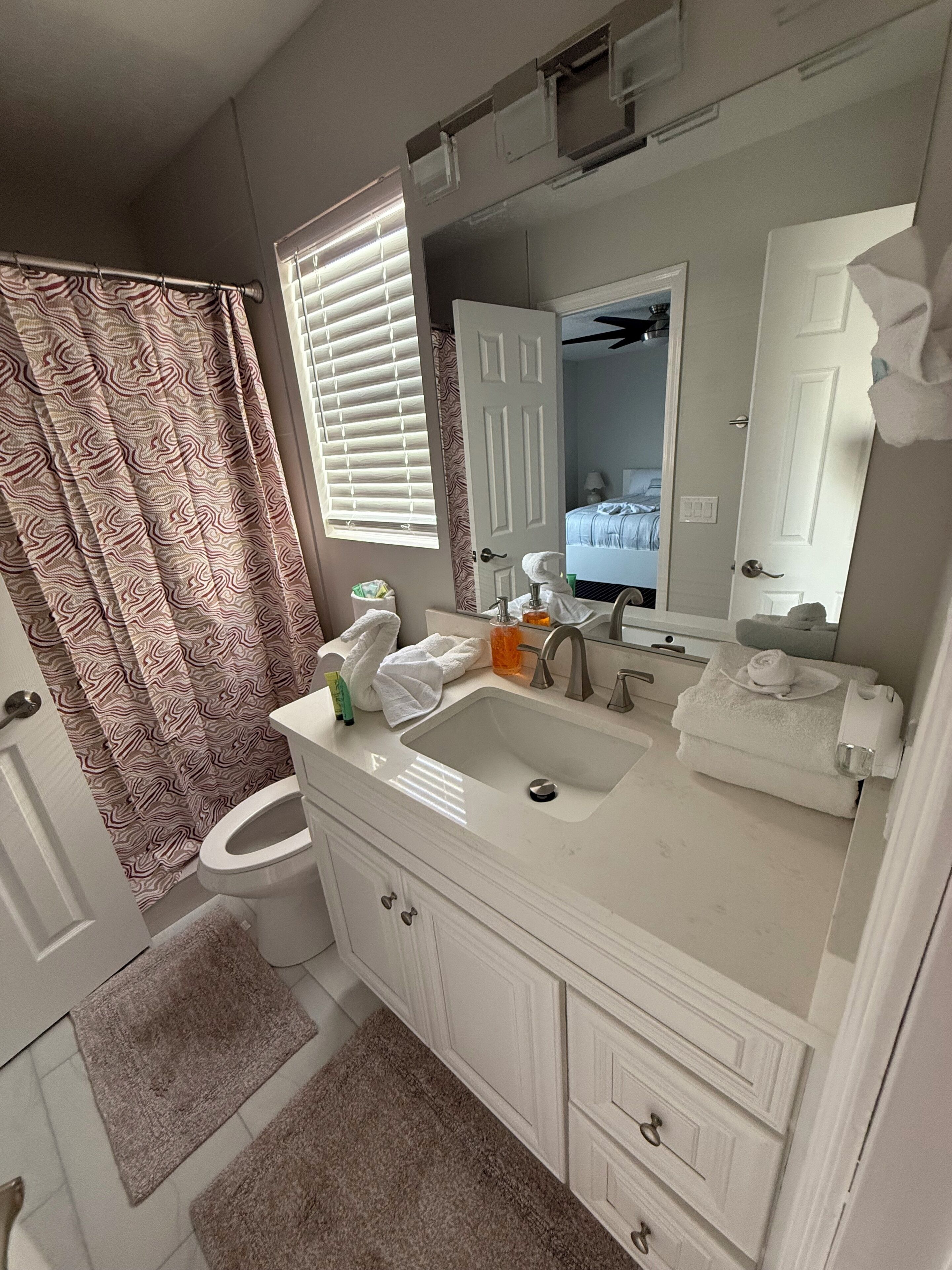 Un bagno con lavandino bianco, specchio e water. Tende da doccia con motivo ondulato.