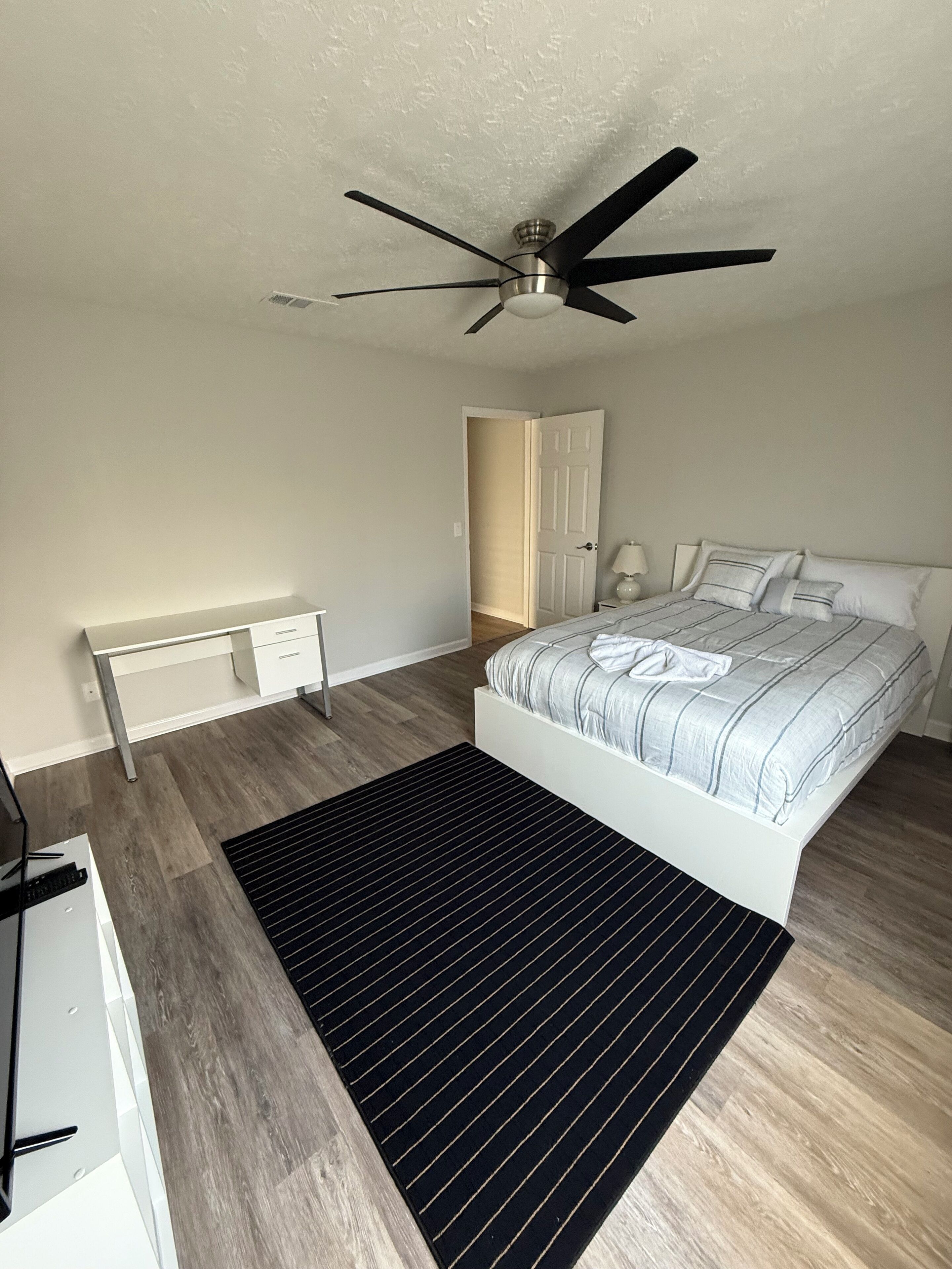A bedroom with a bed, a desk, a chair, a ceiling fan, and a door.