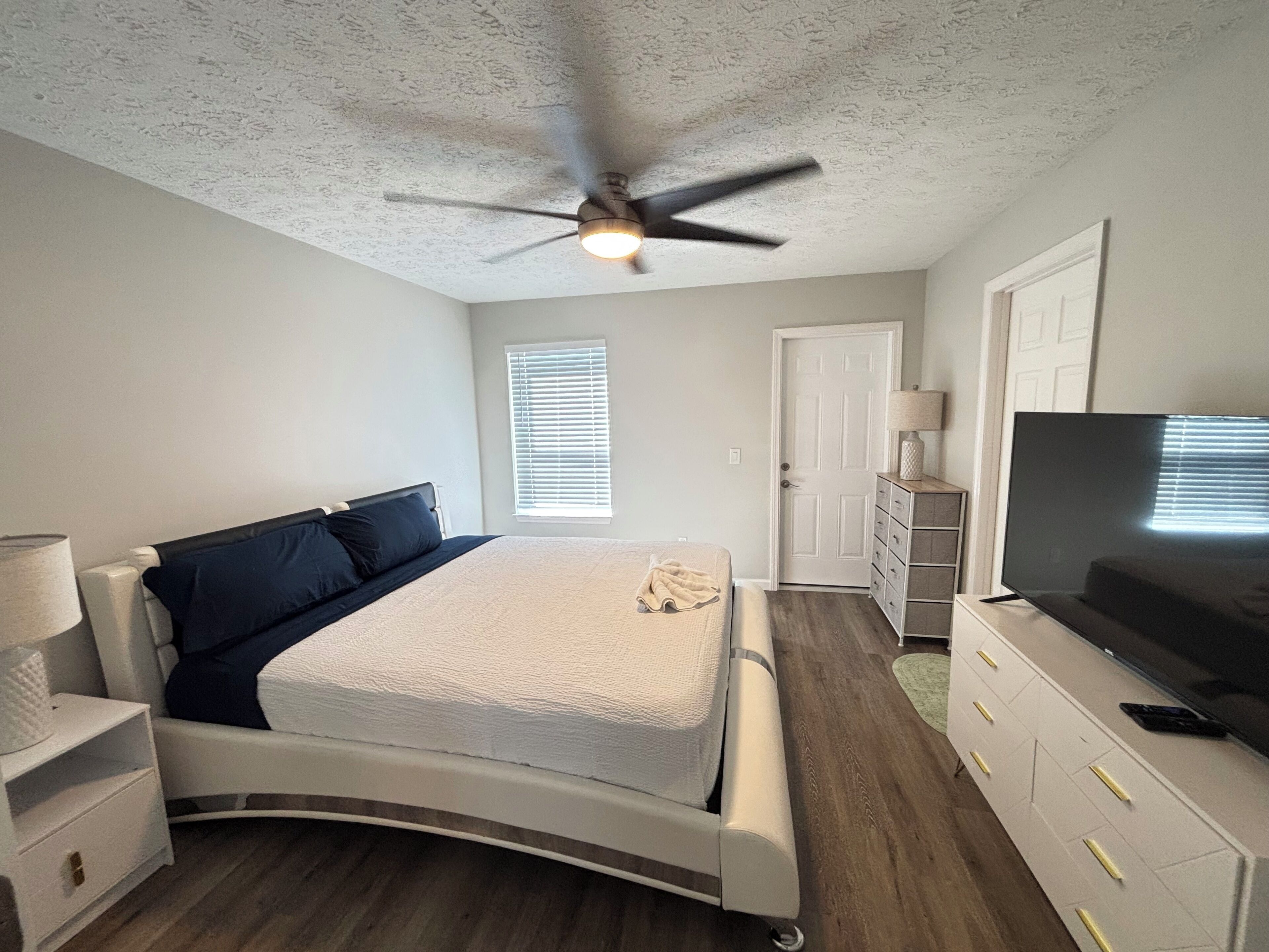 A bedroom with a curved bed, a ceiling fan, a TV, a dresser, and a window with blinds.