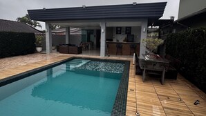 A swimming pool with a tiled deck, a covered patio area, and a view of a residential building in the background.