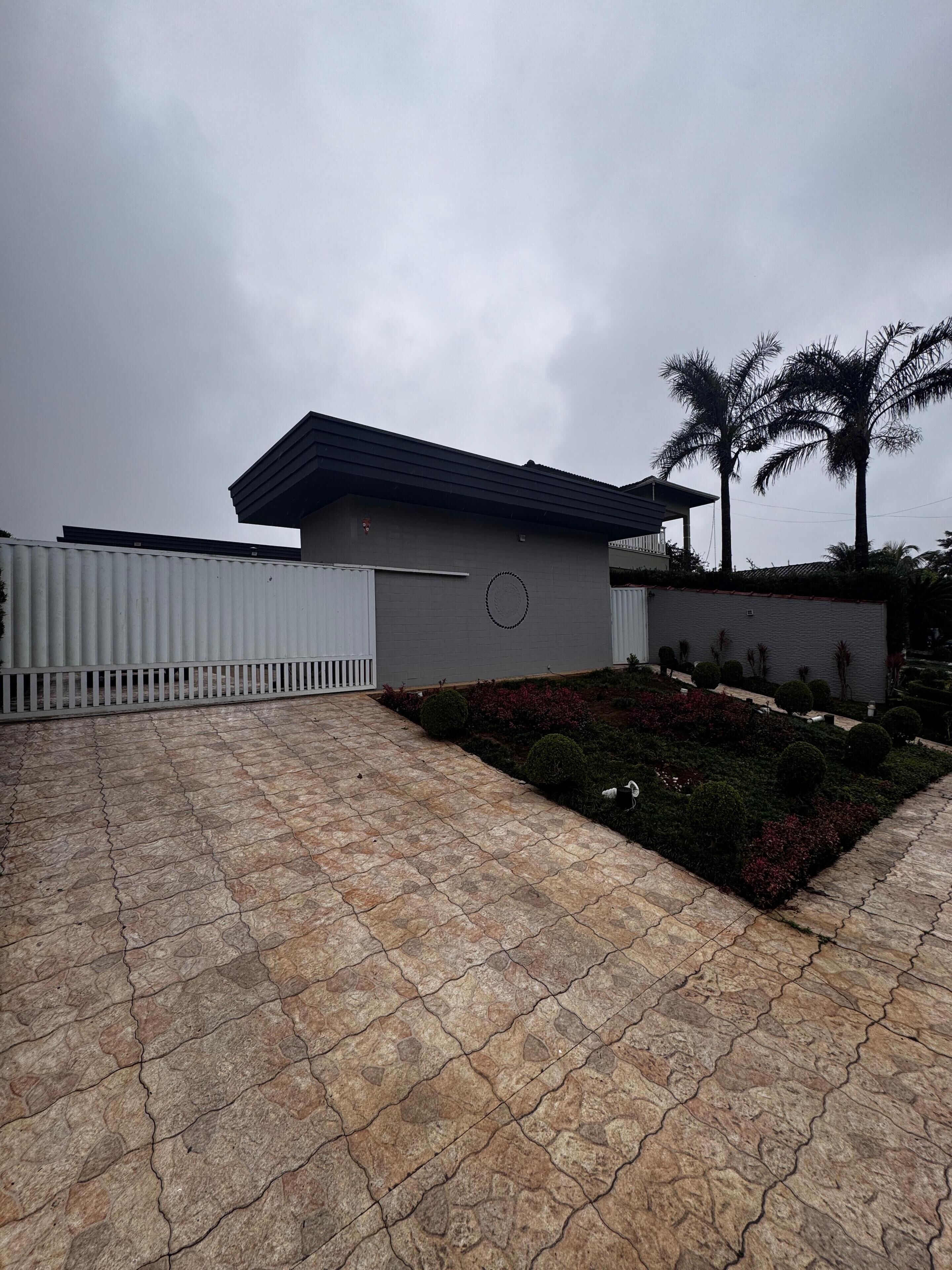 A modern house with a flat roof, a white fence, and a garden with shrubs and palm trees.