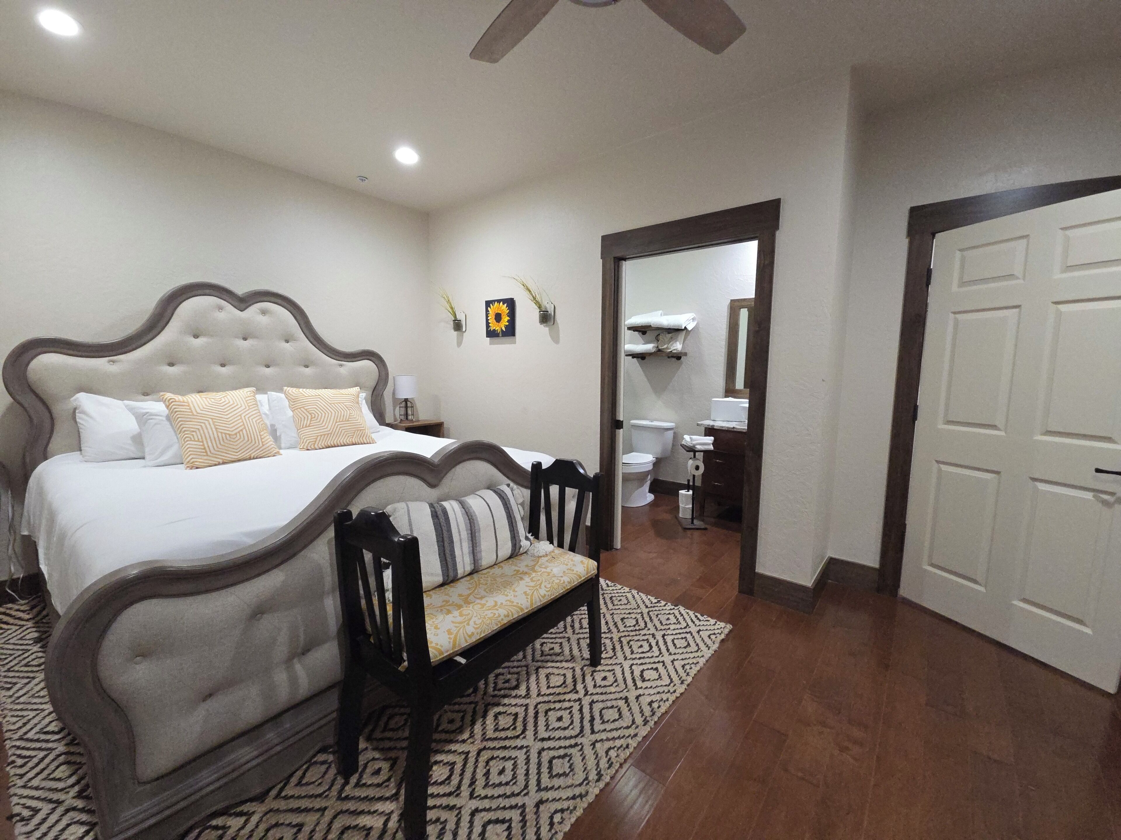 A bedroom with a bed, a bench, a bathroom with a toilet and sink, and a door leading to another room.