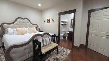 A bedroom with a bed, a bench, a bathroom with a toilet and sink, and a door leading to another room.