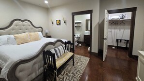 A bedroom with a large bed, bedside table, lamp, and a bench with a patterned rug. - Bluebird Place (Elk City)