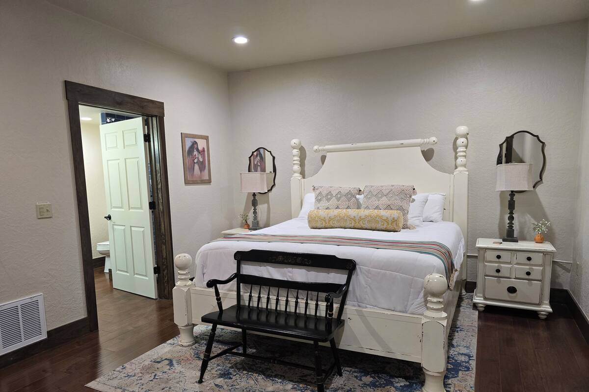 A bedroom with a white four-poster bed, a black chair, a bedside table with a lamp, and a nightstand.
