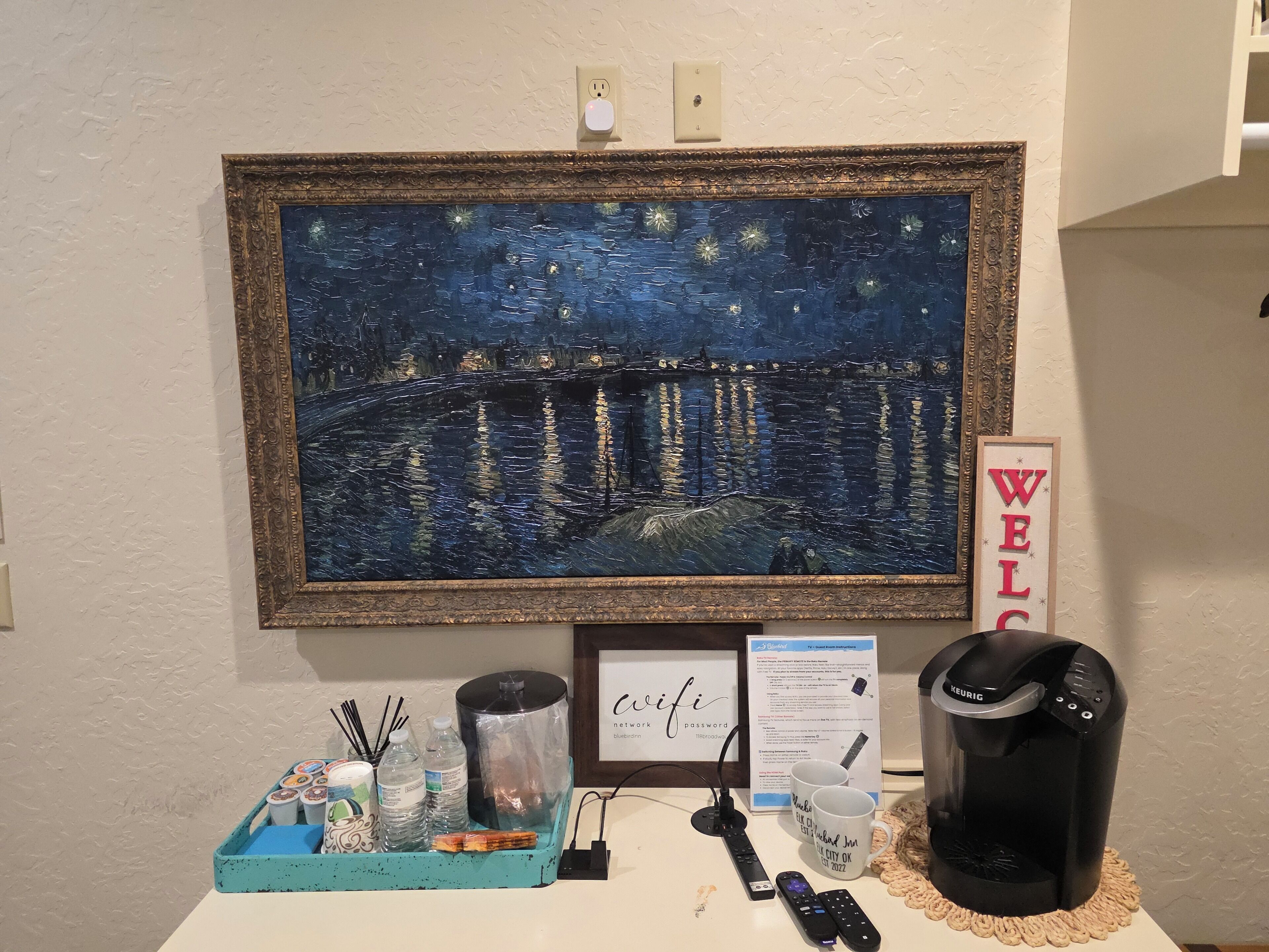 A framed artwork of a night scene, a 'WELCOME' sign, a coffee maker, a remote control, and a sign with Wi-Fi details on a table.