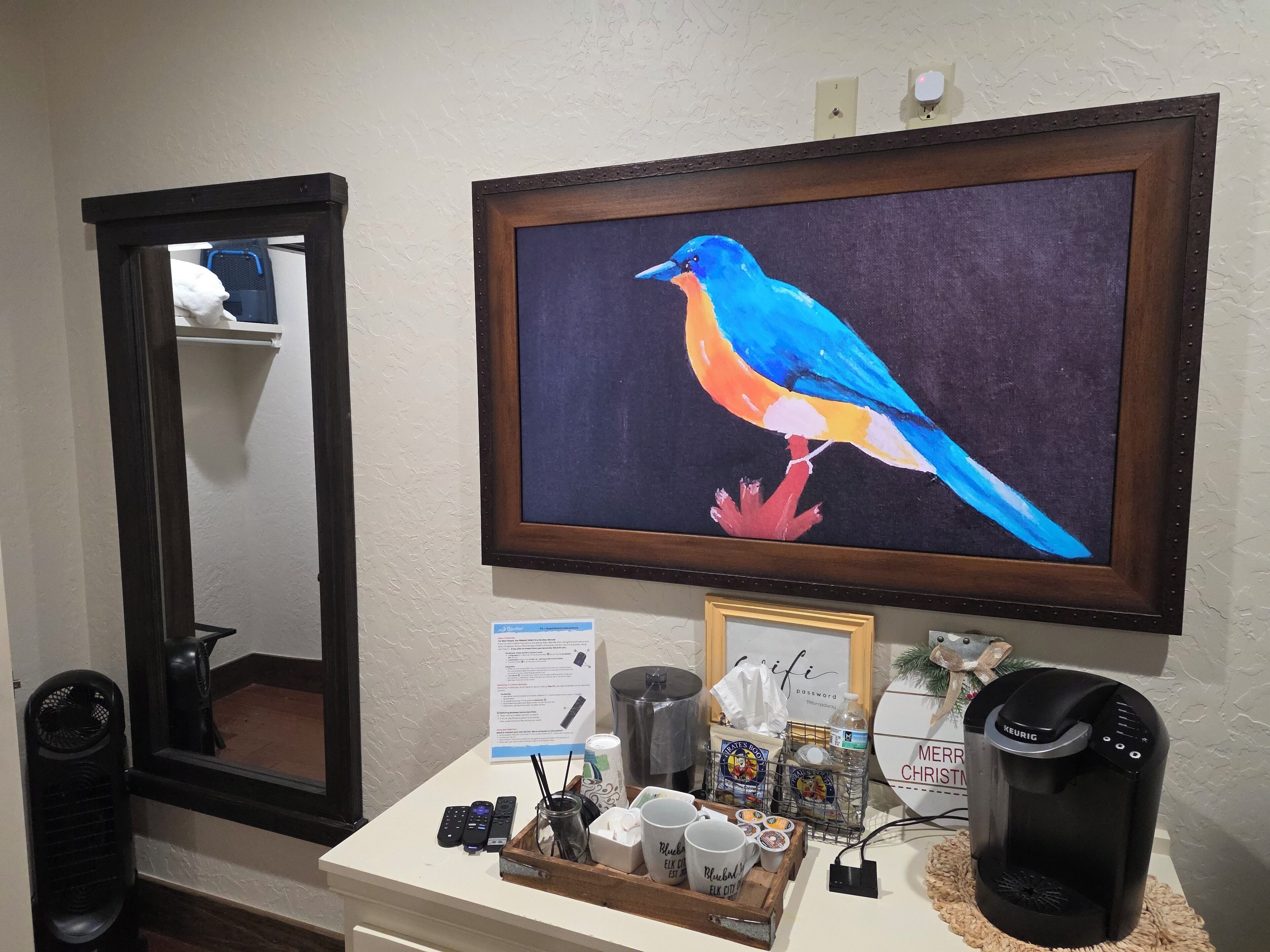 A hotel room with a large mirror, a framed painting of a bird, a coffee maker, and a tray with mugs and remote controls.