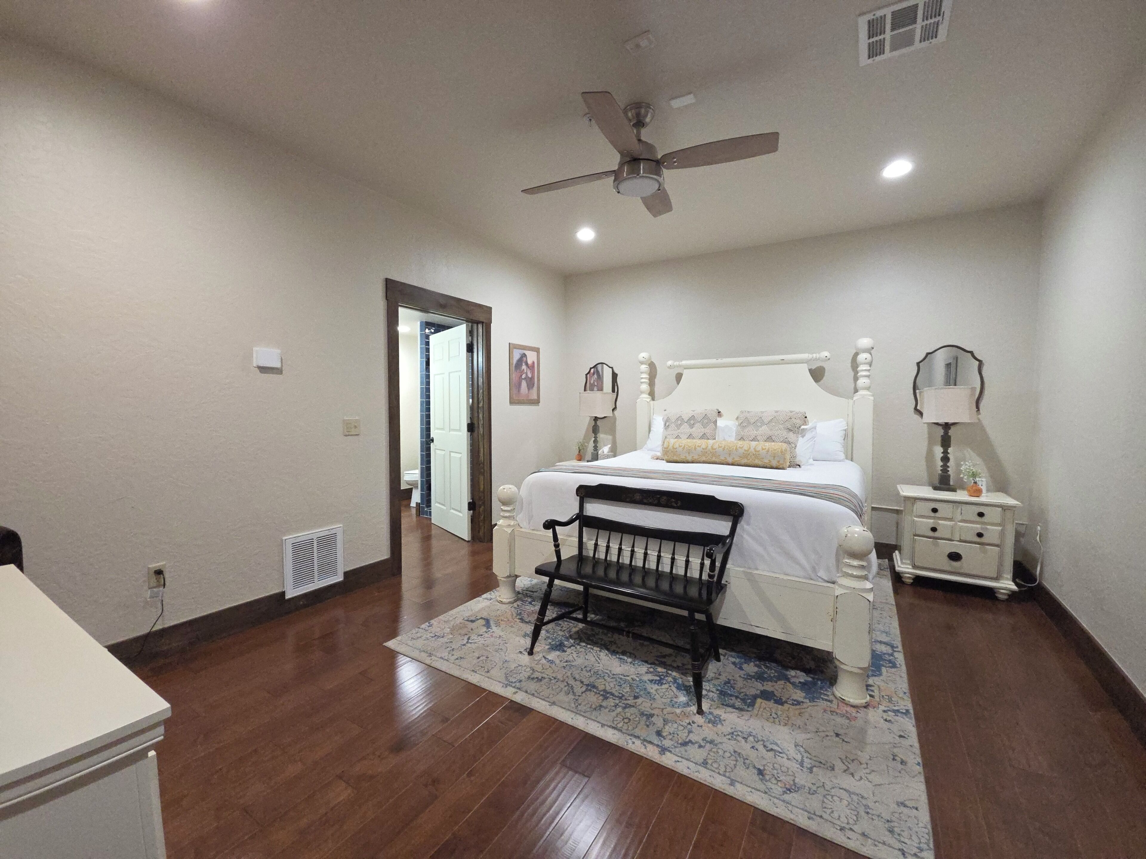 A bedroom with a bed, a chair, a nightstand, a lamp, a ceiling fan, and a door leading to another room.