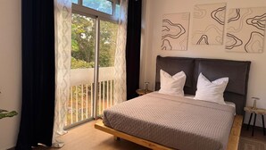 Une chambre à coucher avec un lit, une fenêtre avec des rideaux, un balcon et des œuvres d’art abstraites accrochées au mur.