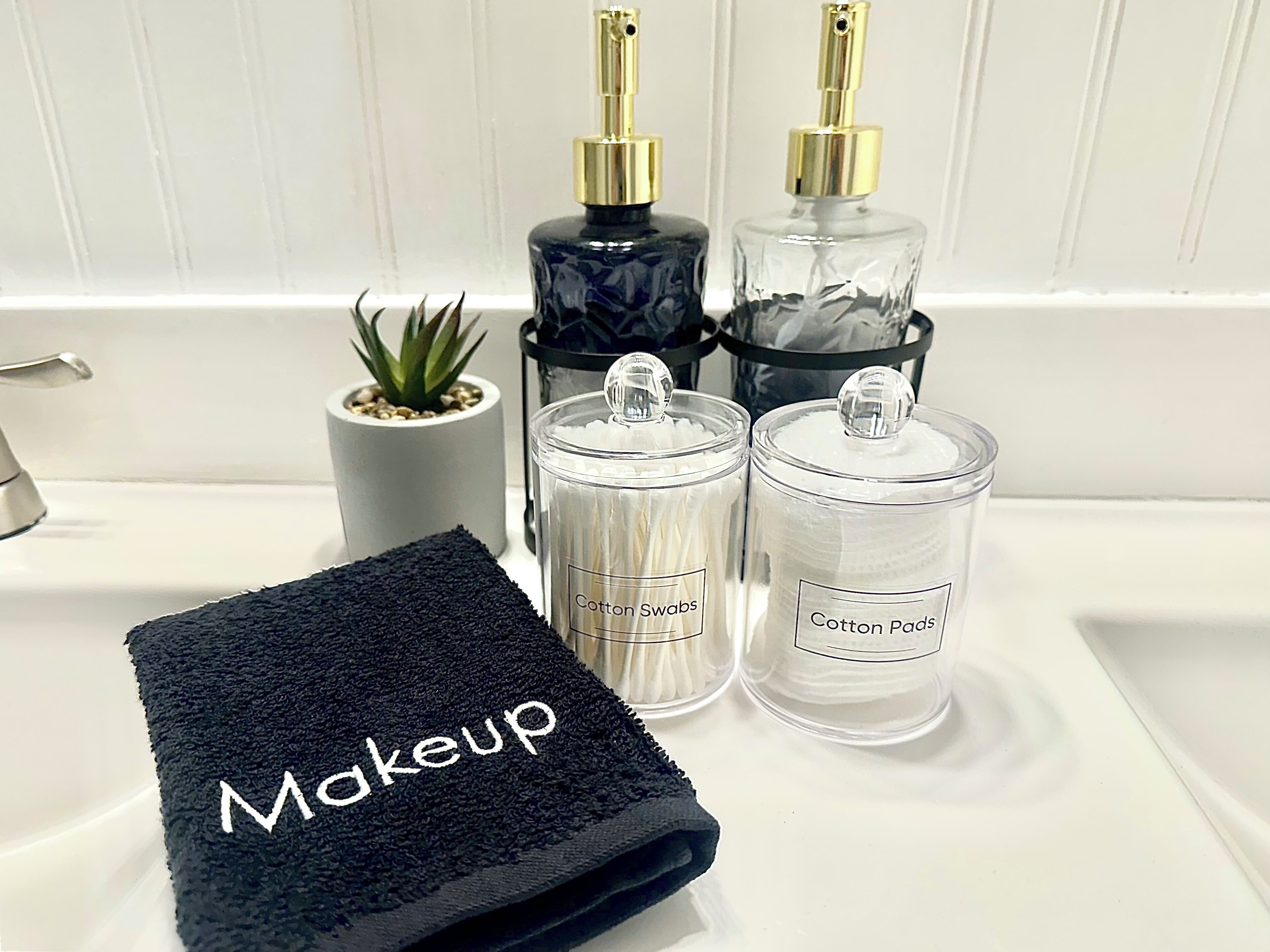 A bathroom sink with a black towel labeled "Makeup," two glass containers labeled "Cotton Swabs" and "Cotton Pads," and a small potted plant.