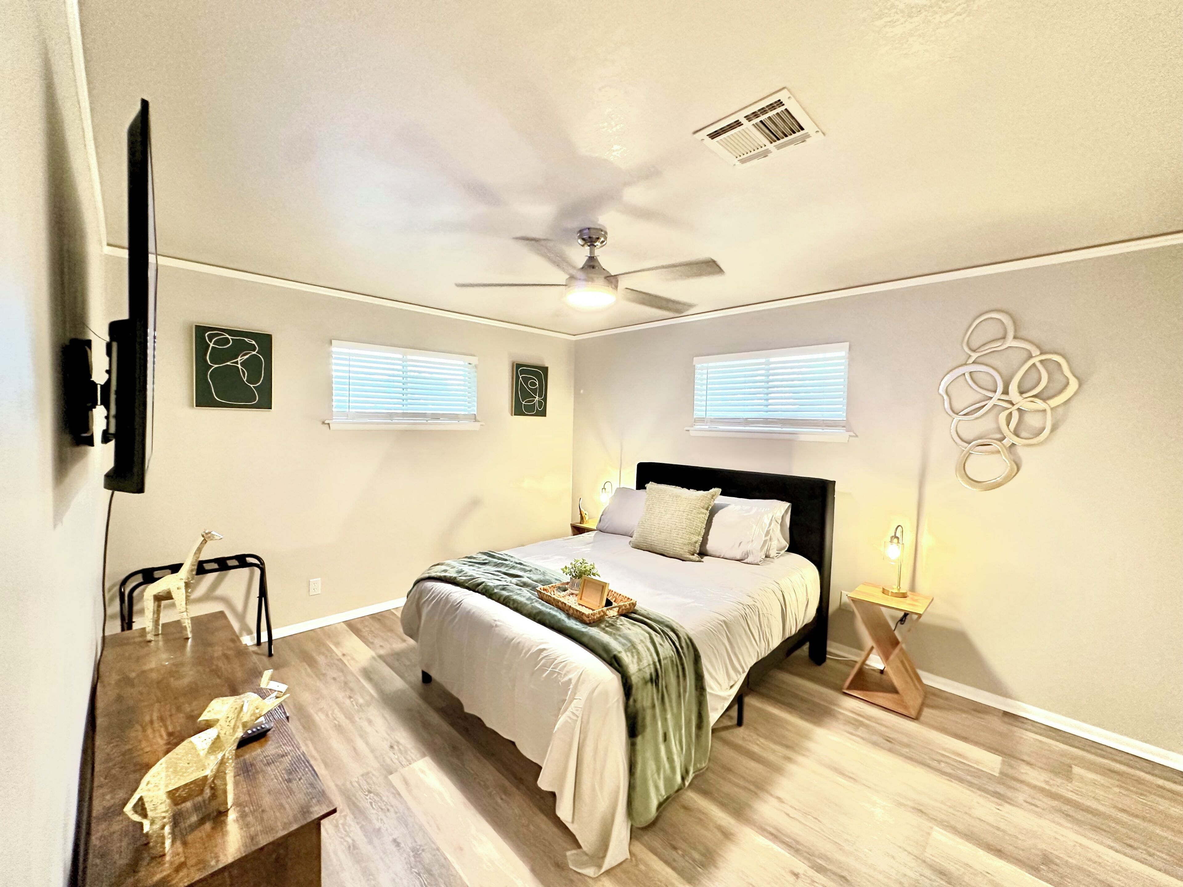 A bedroom with a bed, a ceiling fan, two windows, a wall art piece, and a desk with a chair.