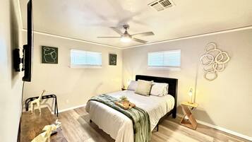 A bedroom with a bed, a ceiling fan, two windows, a wall art piece, and a desk with a chair.