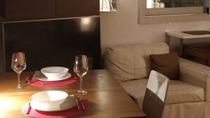 En matplats med ett träbord dukat för två personer, två vinglas och två vita skålar. I bakgrunden står en lädersoffa, och genom ett fönster kan man se ut över byggnaderna.