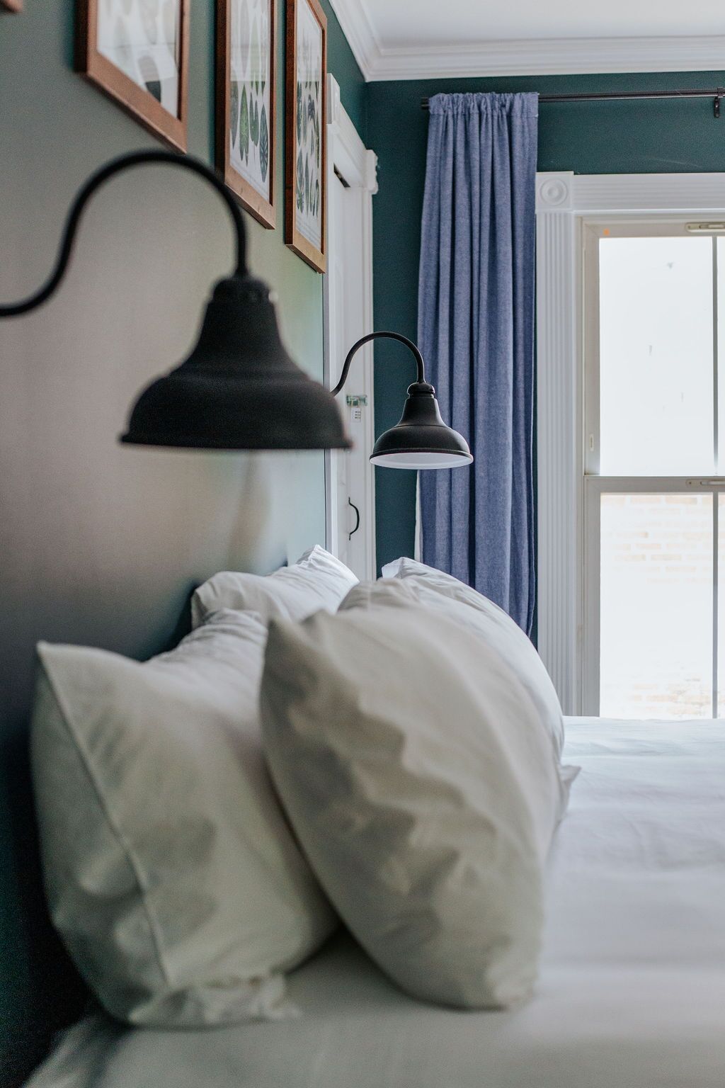 A bedroom with a bed, white pillows, a black wall-mounted lamp, and a window with blue curtains.
