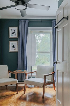 Een kamer met twee stoelen, een tafel, een plafondventilator, een raam met gordijnen en een deur.