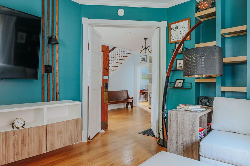 A modern living room with teal walls, a wooden floor, a TV mounted on the wall, and a staircase leading to another room.