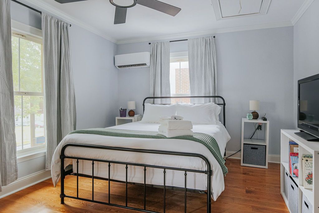 Une chambre avec un lit en métal, du linge de lit blanc, un ventilateur de plafond, un appareil de climatisation, une télévision et une bibliothèque.