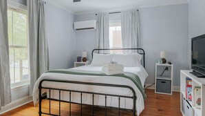 A bedroom with a metal bed frame, white bedding, a ceiling fan, an air conditioning unit, a TV, and a bookshelf.