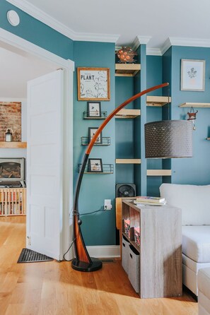 Ein Zimmer mit türkisfarbenen Wänden, einer weißen Tür, Holzboden, einem weißen Sofa, einer Lampe, Regalen mit Dekoration und einem gerahmten Bild an der Wand.