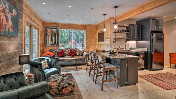 A modern kitchen with wooden accents, a dining area with a sofa and chairs, and a large window offering a view of the outdoors. - Treetop Terrace Cottage (Interlochen)