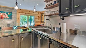 A modern kitchen with stainless steel appliances, wooden cabinetry, and a large window offering a view of the outdoors. - Treetop Terrace Cottage (Interlochen)