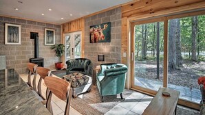 A modern interior with a stone wall, a fireplace, and a large window overlooking a forest. - Treetop Terrace Cottage (Interlochen)
