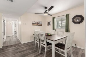 Un coin repas avec une table en bois et des chaises blanches, un ventilateur de plafond, une fenêtre avec des stores et une horloge murale.