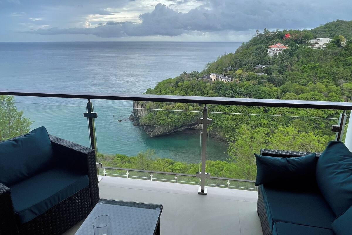 A balcony with wicker furniture and a glass table, overlooking a coastal area with a view of the sea and a hillside with houses.