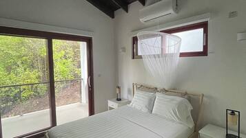 A bedroom with a bed, a ceiling fan, an air conditioning unit, a bedside table, and a window with a mosquito net.