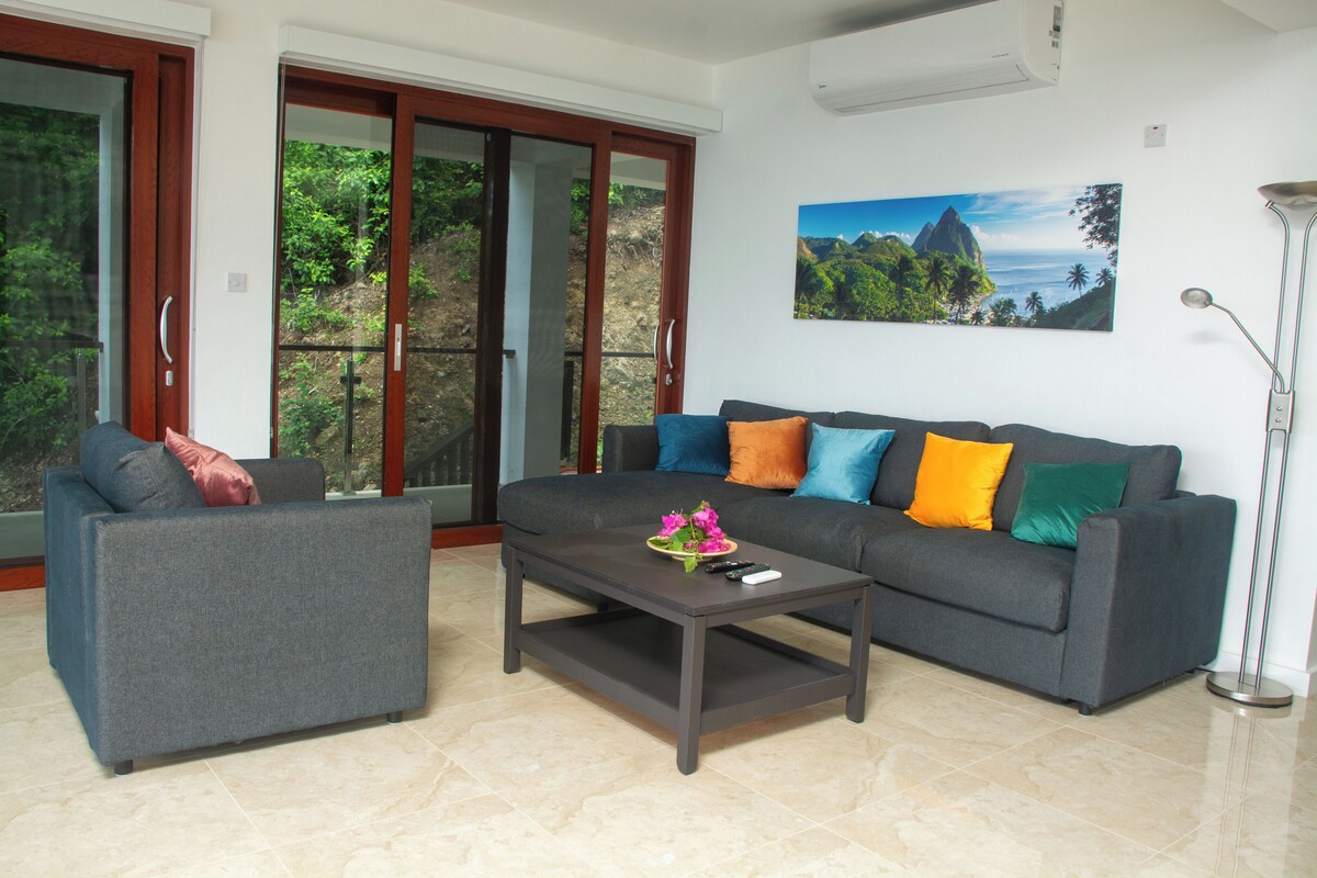 A living room with a grey sofa, a smaller grey armchair, a coffee table, and a painting of a mountainous landscape.
