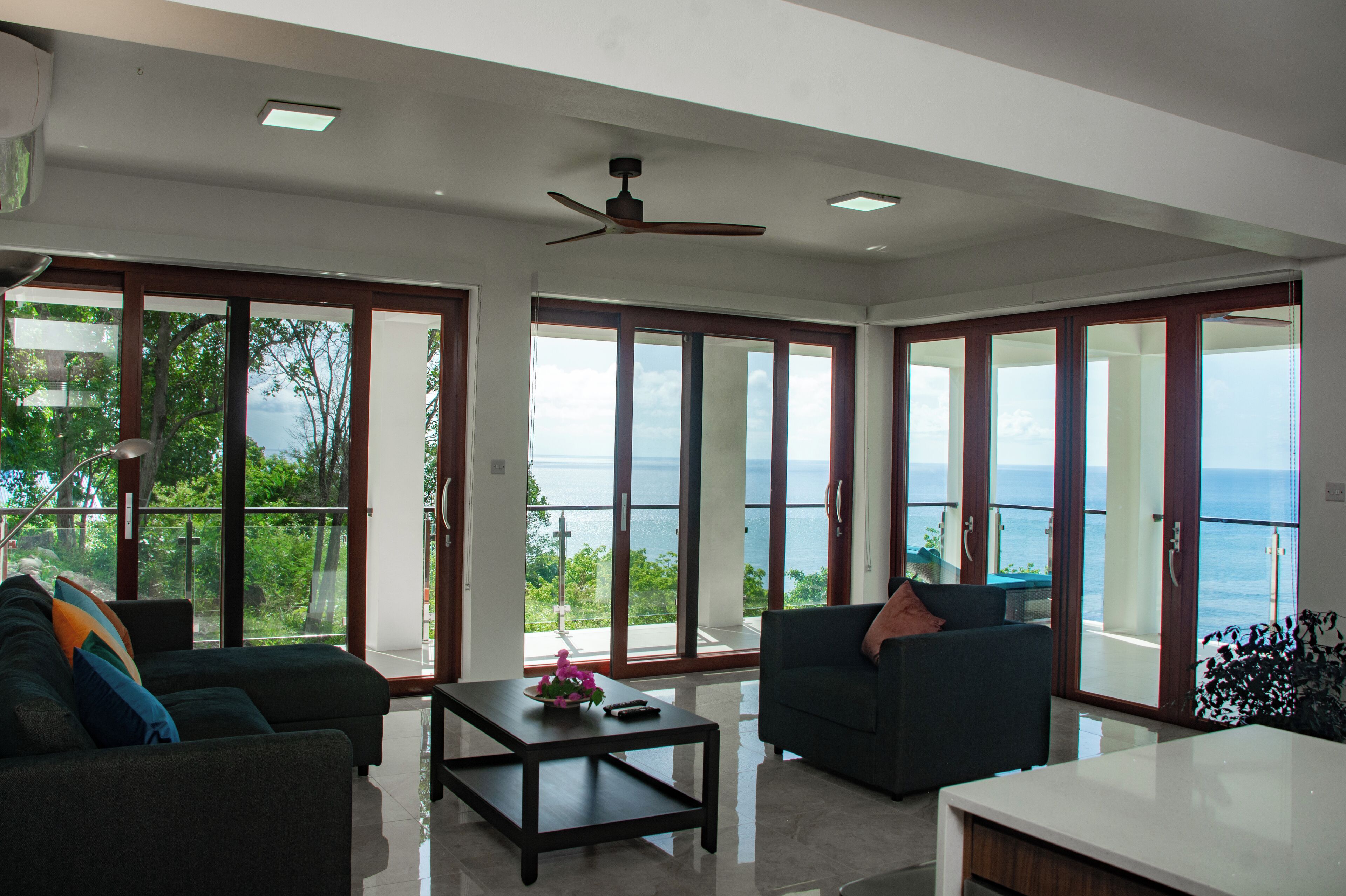 Un salón amplio con un gran ventanal con vista al mar, un ventilador de techo y una decoración interior moderna.