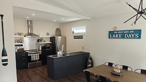 A modern kitchen with a central island, a dining area with a wooden table and chairs, and a sign on the wall that reads "The best days are Lake days." - Turtle Loft Lakefront Cabin Townhome (Kootenay Boundary E / West Bo*)
